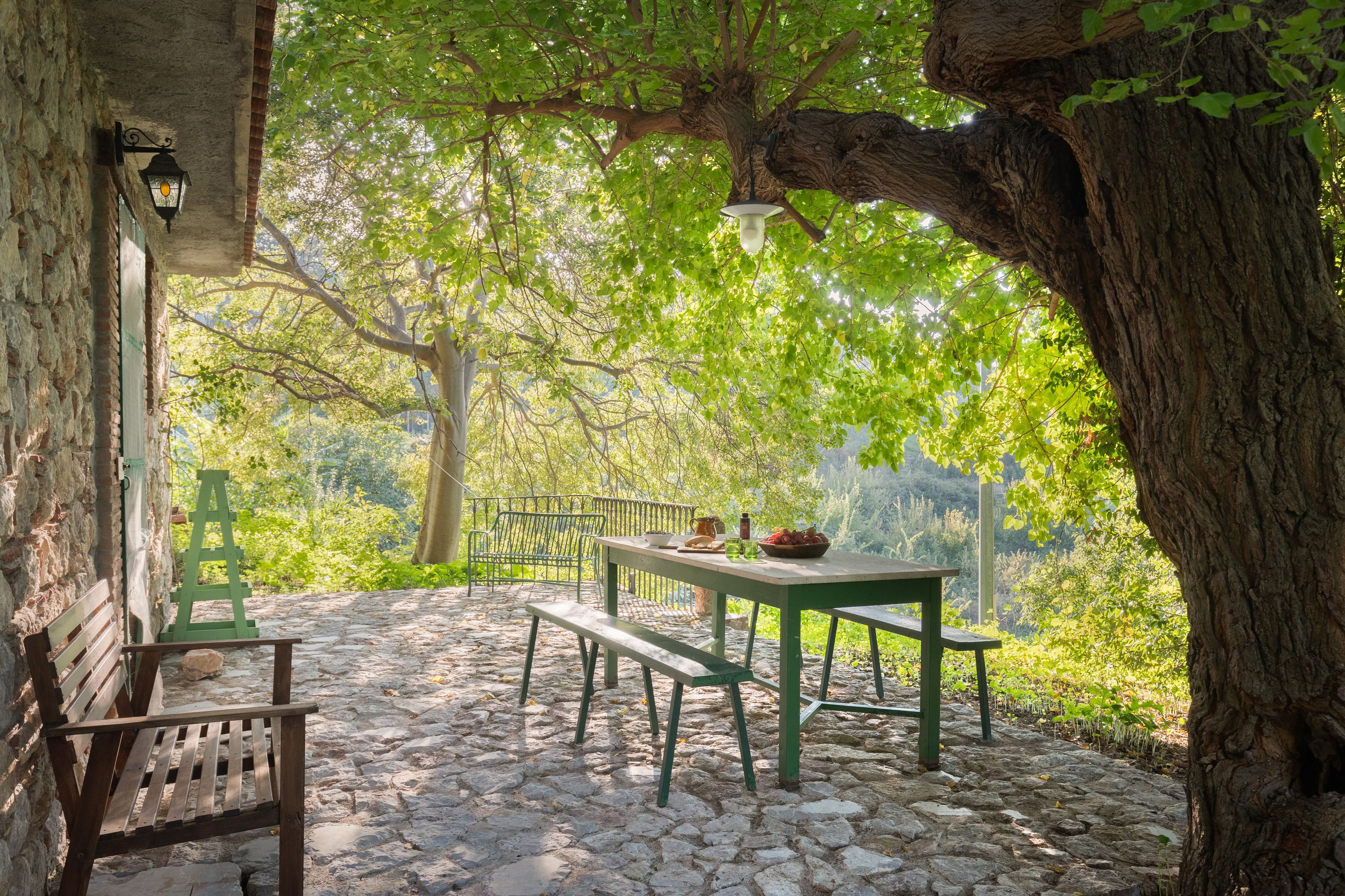 Un espace extérieur aménagé avec un sol en pierre, comprenant une table et des bancs, entouré d’arbres et d’un mur de pierre.