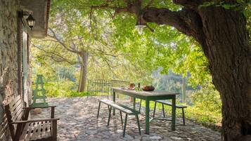 Un espace extérieur aménagé avec un sol en pierre, comprenant une table et des bancs, entouré d’arbres et d’un mur de pierre.