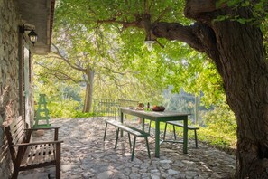 A stone-paved outdoor seating area with a table and benches, surrounded by trees and a stone wall.