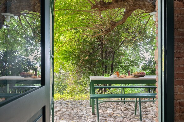 A stone patio with a table and bench, surrounded by lush greenery and a tree.