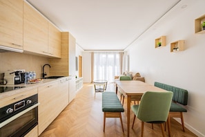 A modern kitchen with wooden cabinets, a dining area with a wooden table and green upholstered chairs, and a view of the city through large windows. - Saint Denis—Metro—3 Bedrooms—Modern—New (Saint-Denis)