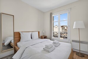 A hotel room with a bed, a nightstand, a lamp, a window with a view, and a radiator. - Flexible Lease – Modern – 1 Bedroom – Parisian Charm (Levallois-Perret)