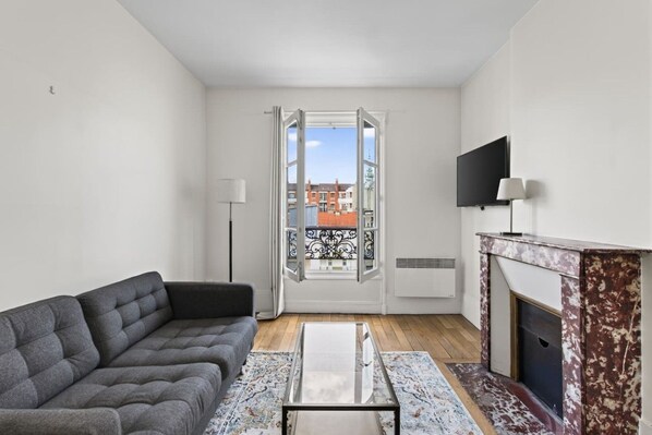 A modern living room with a grey sofa, a fireplace, a TV mounted on the wall, and a large window offering a view of the city. - Flexible Lease – Modern – 1 Bedroom – Parisian Charm (Levallois-Perret)