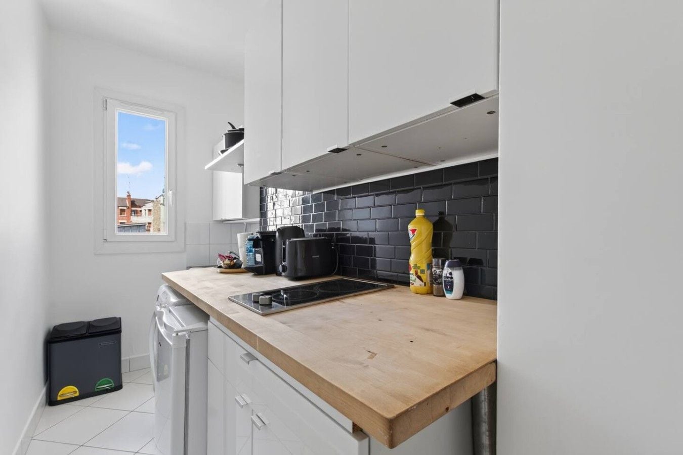 A modern kitchen with a black tiled backsplash, a wooden countertop, and a window with a view of buildings.