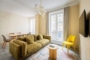 Ein Wohnzimmer mit einer Couch, einem Esstisch, Stühlen, einem Couchtisch, einem Fernseher und einem Fenster mit Vorhängen.