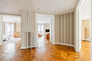 An open-plan living space with a herringbone wooden floor, a radiator, and a view through an open door to another room. - Rome-Premium-Parc Monceau-Villiers-3-Bedroom-Haussmann-Style (Paris)
