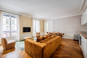 A living room with a large sofa, a television, a dining table, and a kitchen area. - Rome-Premium-Parc Monceau-Villiers-3-Bedroom-Haussmann-Style (Paris)