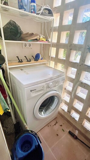 A compact laundry room with a front-loading washing machine, shelves with various items, and a window with a grid pattern. - Luxury modern apartment with plunge pool on golf resort (Fuente Alamo)