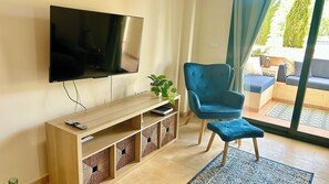 A modern living room with a flat-screen TV mounted on the wall, a wooden entertainment unit with storage, a blue armchair, and a small table with a plant. - Luxury modern apartment with plunge pool on golf resort (Fuente Alamo)