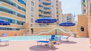 A pool area with lounge chairs and umbrellas, surrounded by multi-story buildings with balconies. - Grazioso Bilocale con Piscina e Terrazza Wi-fi AC (Rome)