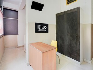 A hotel reception area with a wooden desk, a yellow chair, a black sign indicating the hotel name, and a large dark-colored door.