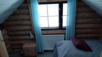 A small, cozy room with a sloped ceiling, a window with blinds, a bedside table with a lamp, and two beds with pillows.