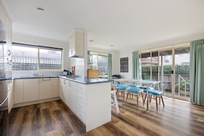 A modern kitchen with a central island, a dining area with a glass table and chairs, and a view of a residential area through large windows. - Bimble Street Beach House - 300 Meters to beach & Pet Friendly (Melbourne)