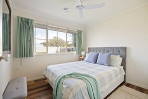 A bedroom with a bed, a ceiling fan, a window with curtains, and a view of the outside. - Bimble Street Beach House - 300 Meters to beach & Pet Friendly (Melbourne)