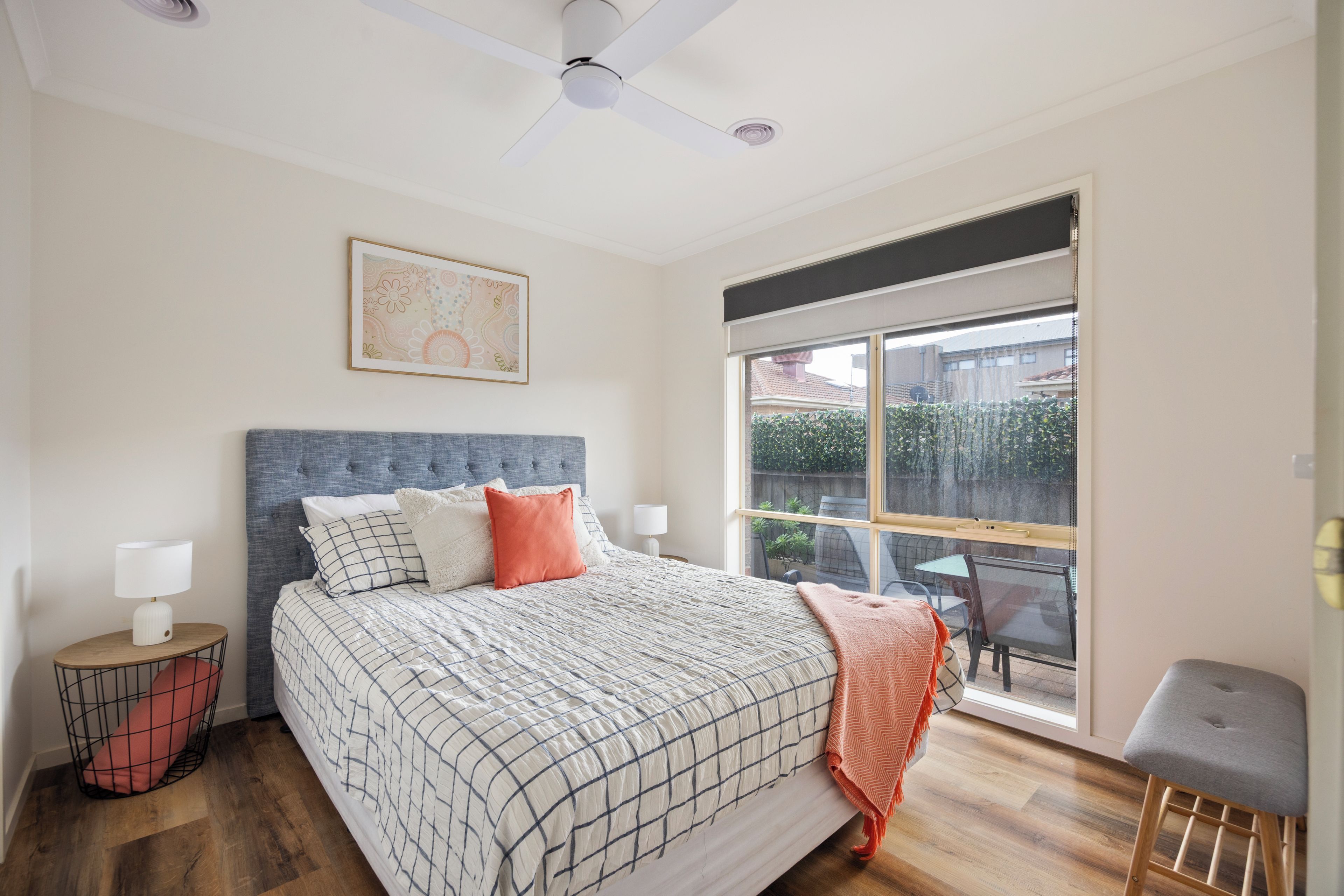 A bedroom with a large bed, a ceiling fan, a window with a view of the outside, and a small table with a lamp.