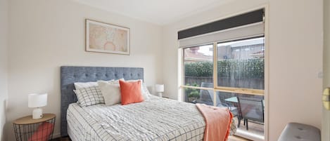A bedroom with a large bed, a ceiling fan, a window with a view of the outside, and a small table with a lamp.
