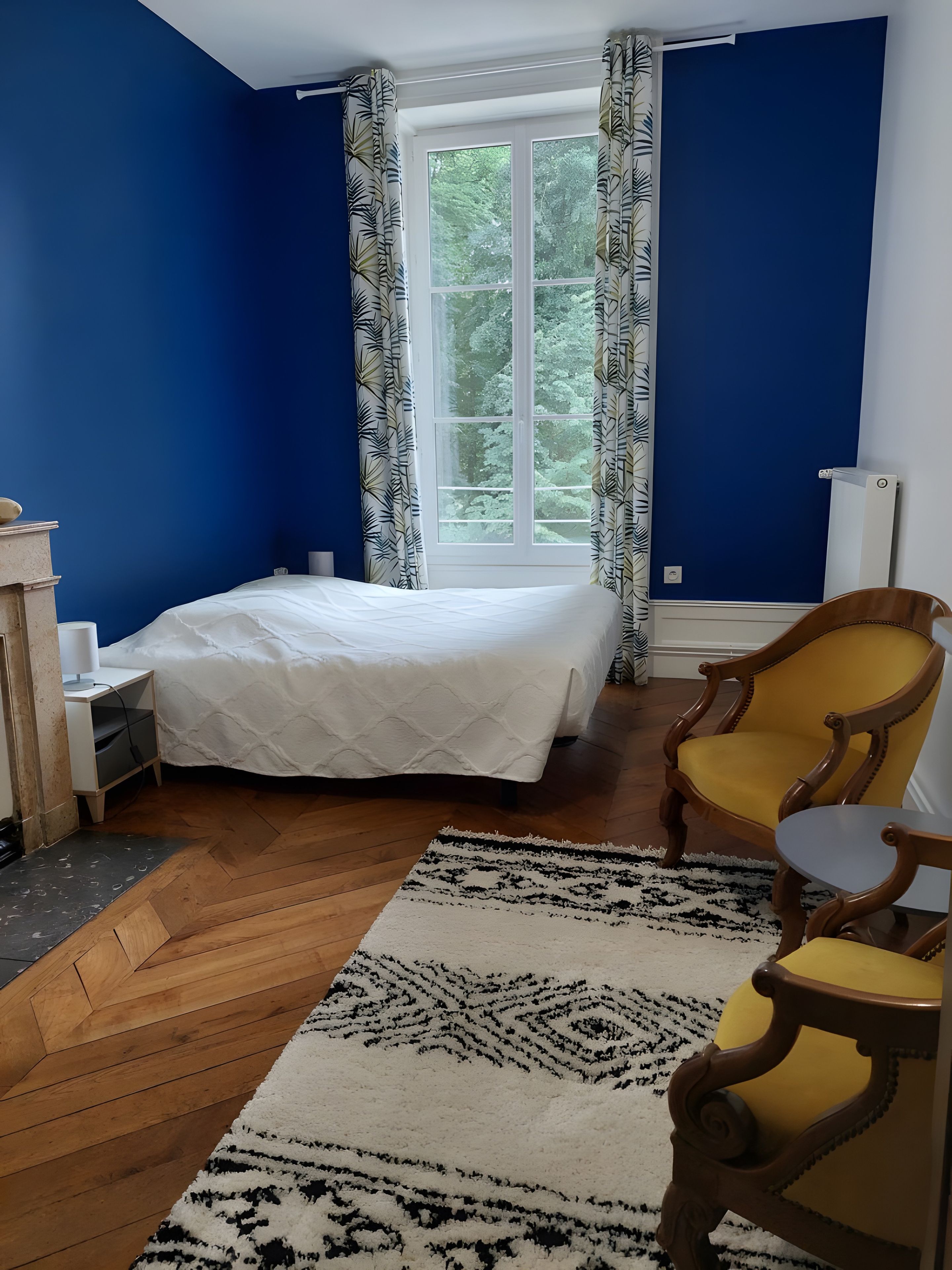 Une chambre à coucher avec un lit, une chaise, une table de chevet, une lampe et une fenêtre avec des rideaux.