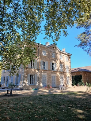 A two-story building with a symmetrical facade, multiple windows with shutters, and a paved courtyard with outdoor seating.
