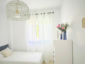 Een slaapkamer met een bed, een witte dressoir, een vaas met bloemen en een wit gordijn.