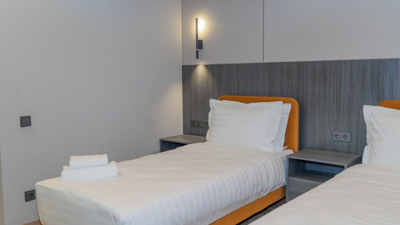 Ein Hotelzimmer mit zwei Betten, jeweils mit weißer Bettwäsche und orangefarbenen Kopfteilen. Über den Betten befindet sich eine Wandleuchte.