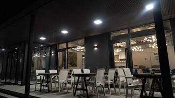 Un área de estar al aire libre con mesas y sillas, un ventanal grande e iluminación empotrada en el techo.