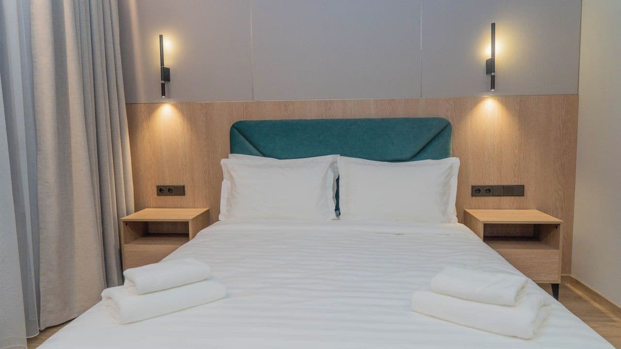 Una habitación de hotel con una cama, dos mesitas de noche, cabecero e iluminación en la pared.