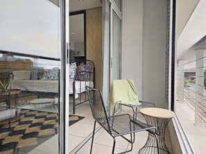 A balcony with a bed, a chair, a small table, and a view of the street. - Modern Apartment Secure and Spacious (Cape Town)