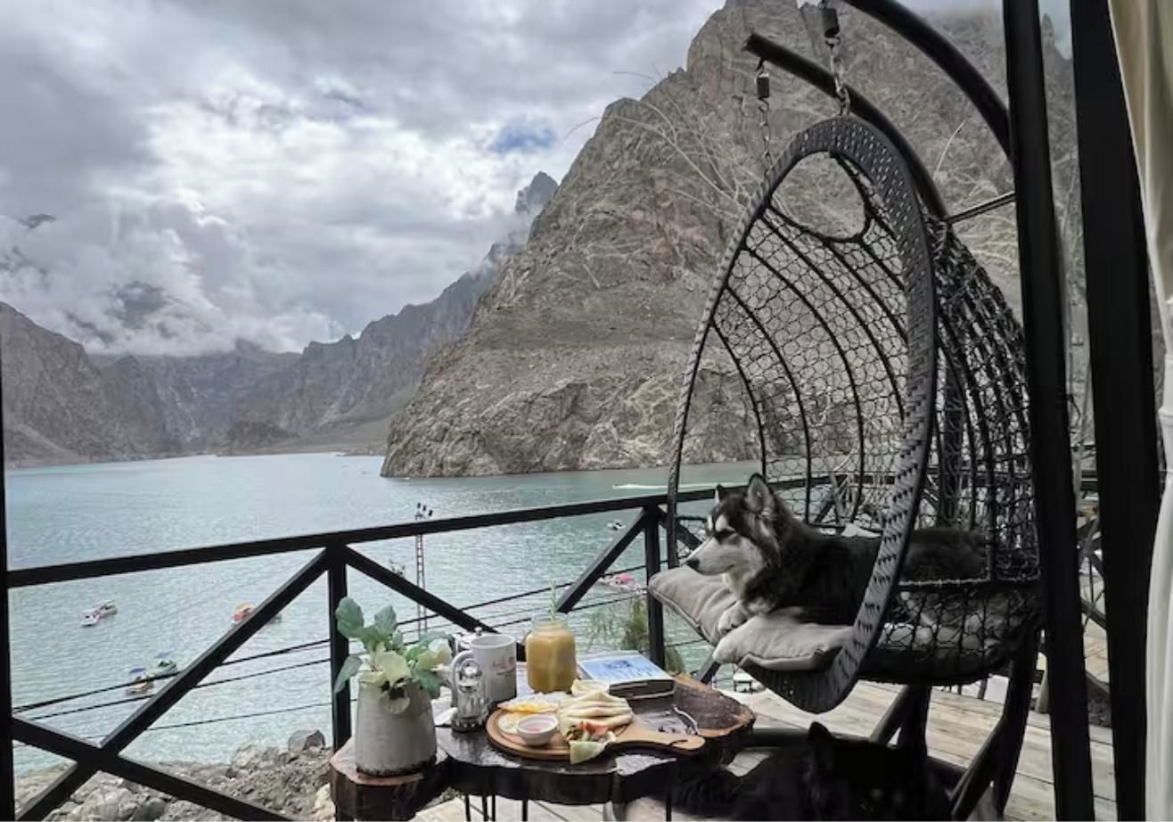 A room with a view of a lake and mountains, a hanging chair with a dog, and a table with food and drinks.