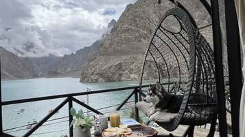 A room with a view of a lake and mountains, a hanging chair with a dog, and a table with food and drinks.