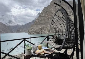 Una habitación con vistas a un lago y montañas, una silla colgante con un perro, y una mesa con comida y bebidas.