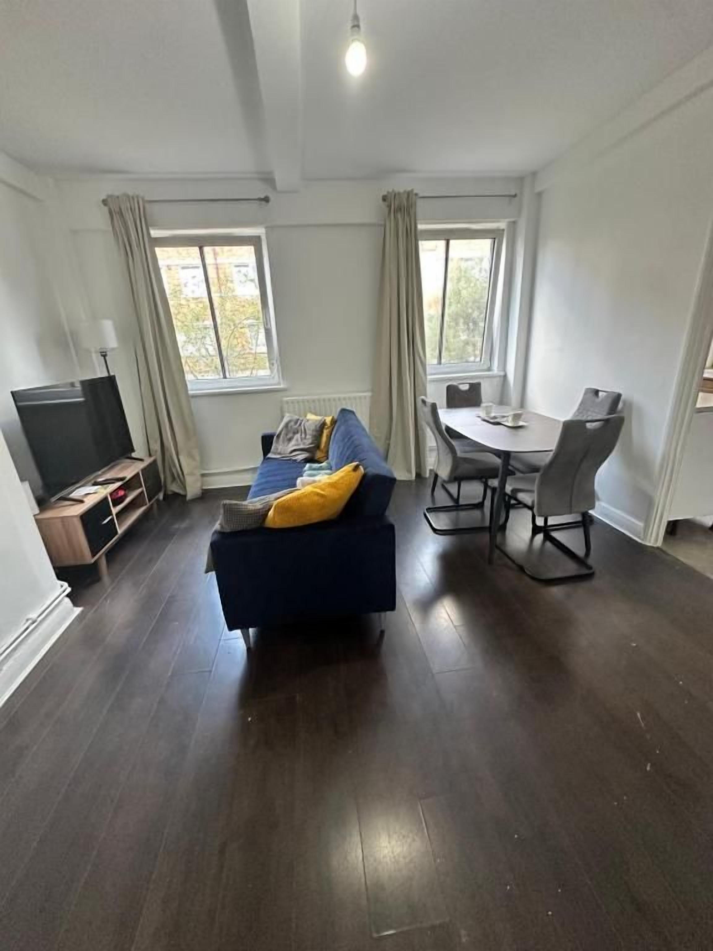 A modern living room with a dark wood floor, a blue sofa with yellow cushions, a dining table with chairs, and a flat-screen TV.