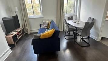 A modern living room with a dark wood floor, a blue sofa with yellow cushions, a dining table with chairs, and a flat-screen TV.