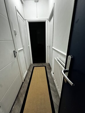 Un couloir étroit, avec des portes blanches de chaque côté, un intérieur sombre visible à travers la porte ouverte, et un radiateur sur le mur de droite.