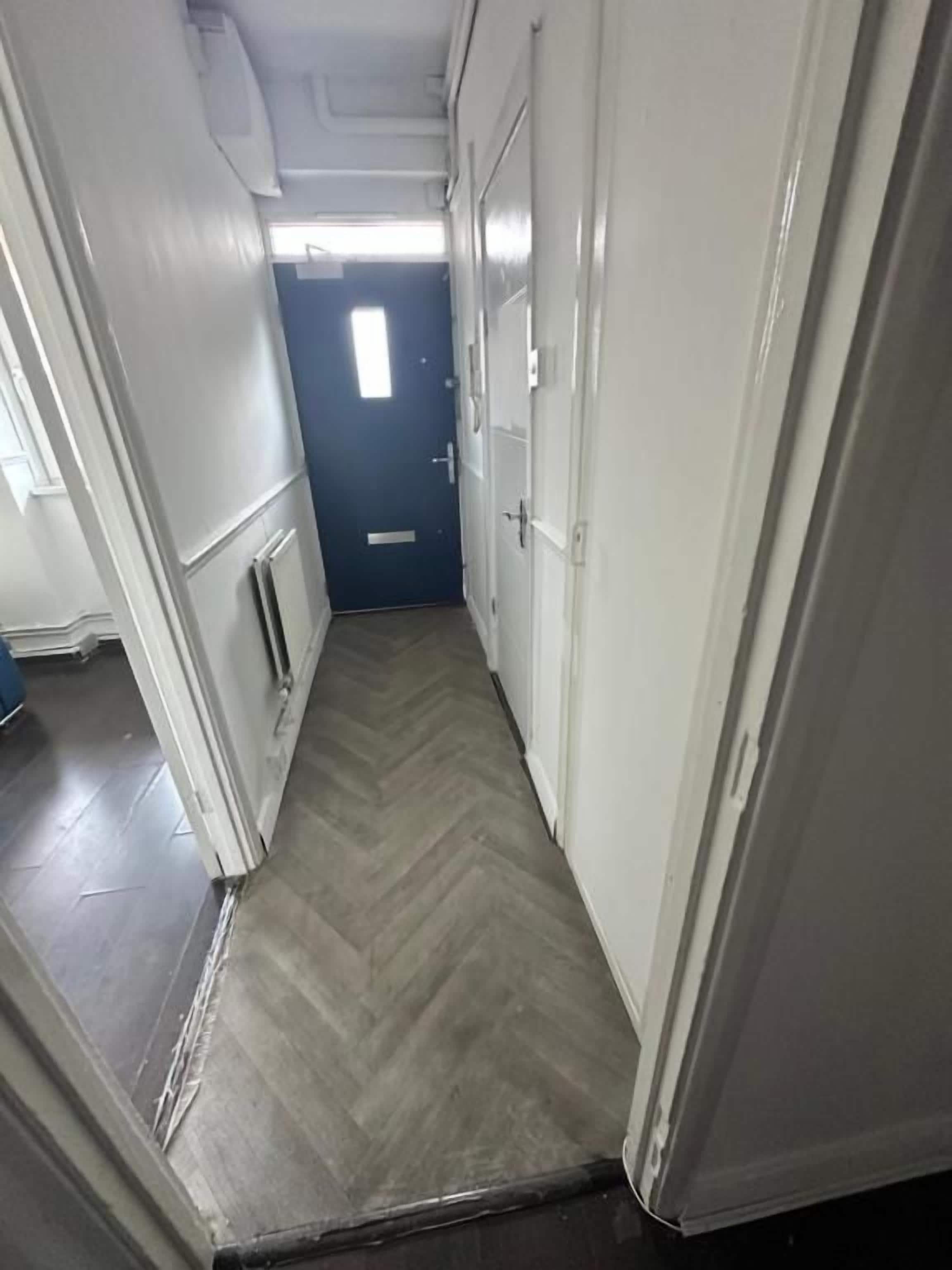 A narrow hallway with a herringbone floor pattern, white walls, and a door at the end.