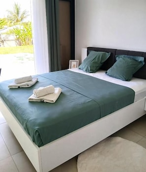 Una cama con colcha verde y dos almohadas, una mesita de noche blanca con un cuadro enmarcado, y vista a zonas verdes al exterior.