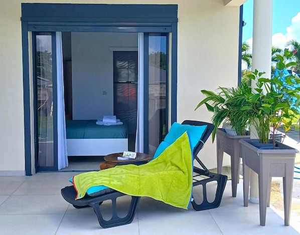Un balcón con una silla de descanso, una planta en maceta y una puerta corrediza de vidrio que da a un dormitorio.