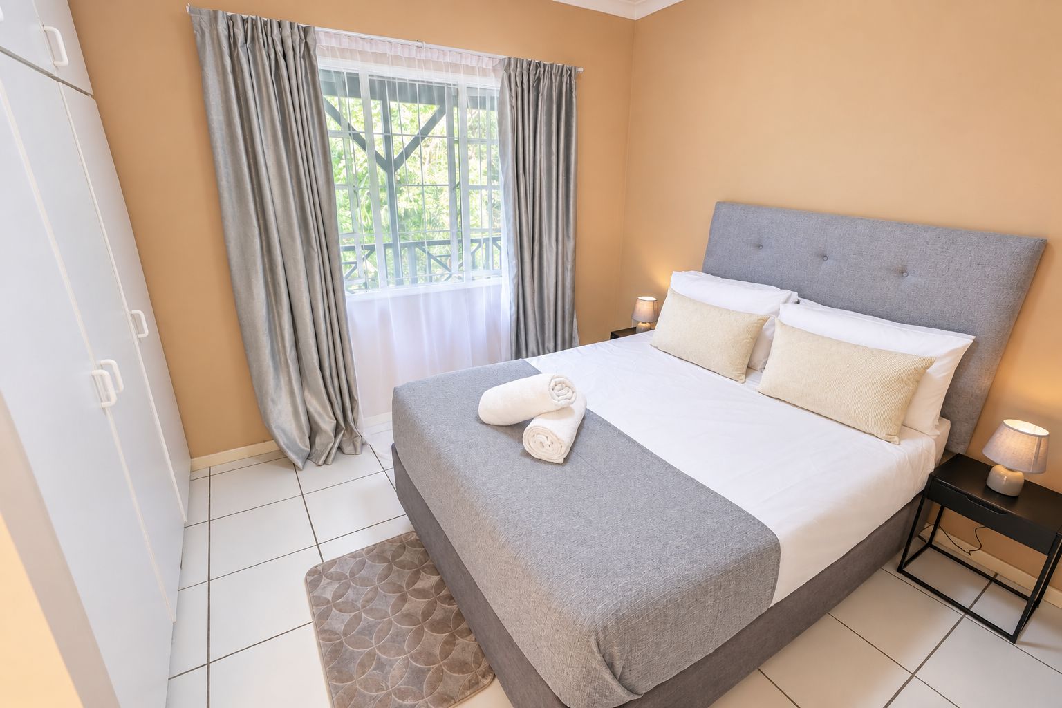 Una cama bien hecha con almohadas y toallas enrolladas, una mesita de noche con lámpara, una ventana con cortinas y una puerta con pomo.