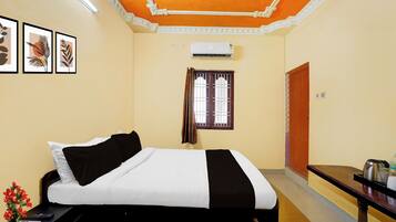A hotel room with a bed, a window with curtains, a wall-mounted air conditioner, and a framed picture on the wall.