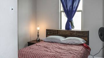 A bedroom with a bed, a nightstand with a lamp, a window with curtains, and a wall-mounted fan.