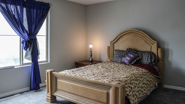Een slaapkamer met een houten bed, een nachtkastje, een lamp en een raam met blauwe gordijnen.