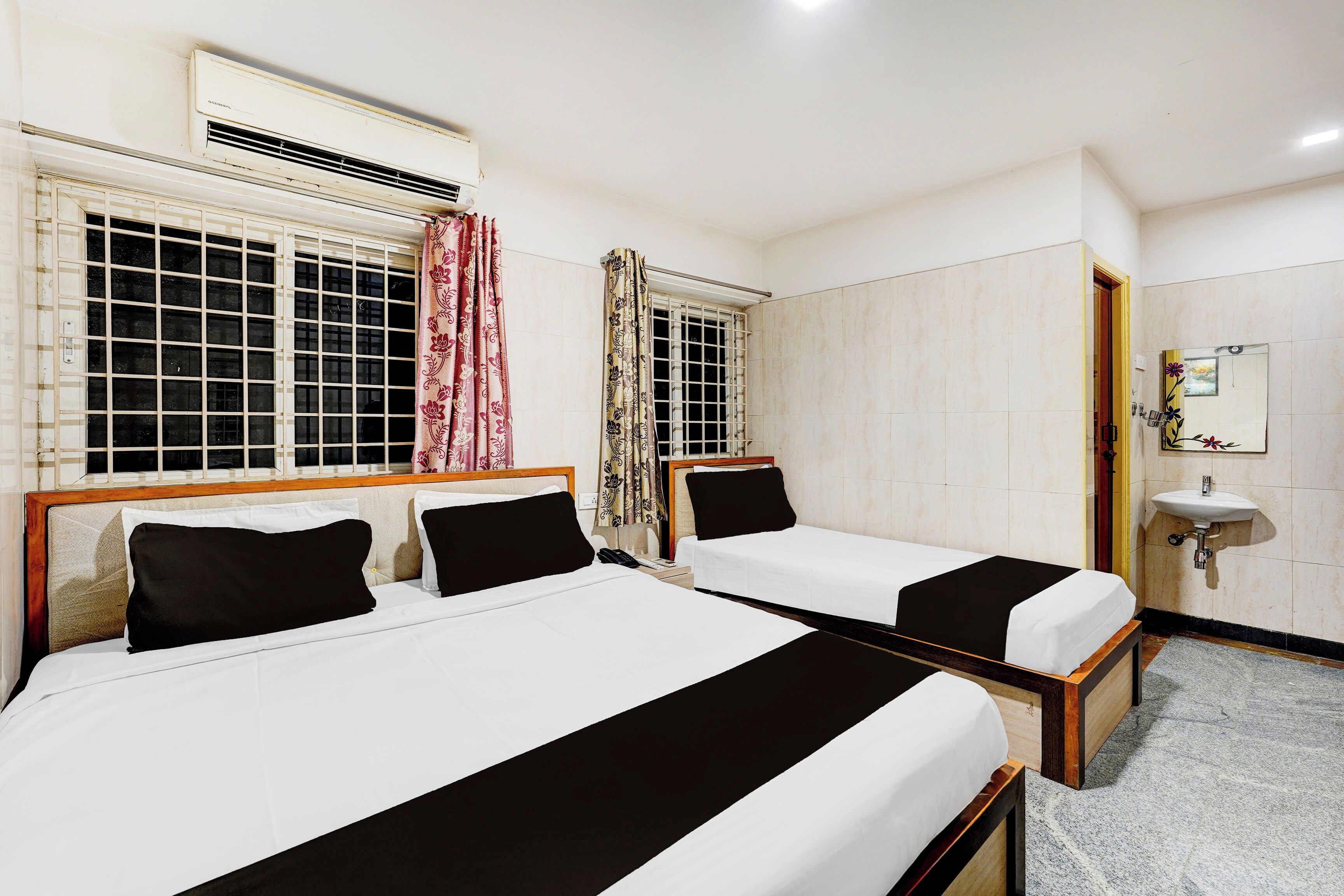 A hotel room with two single beds, a window with a curtain, a bedside table, and a wall-mounted sink.