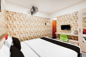 Hotellihuone, jossa on sänky, työpöytä tuolilla, televisio ja seinään kiinnitetty tuuletin.