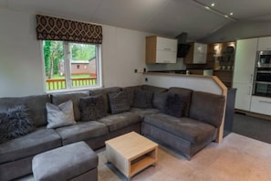A modern living room with a grey sectional sofa, a wooden coffee table, and a view of a backyard through a window with patterned curtains.