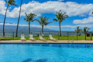Ein Pool mit Liegestühlen und Palmen, mit Blick auf das Meer und die Berge im Hintergrund.