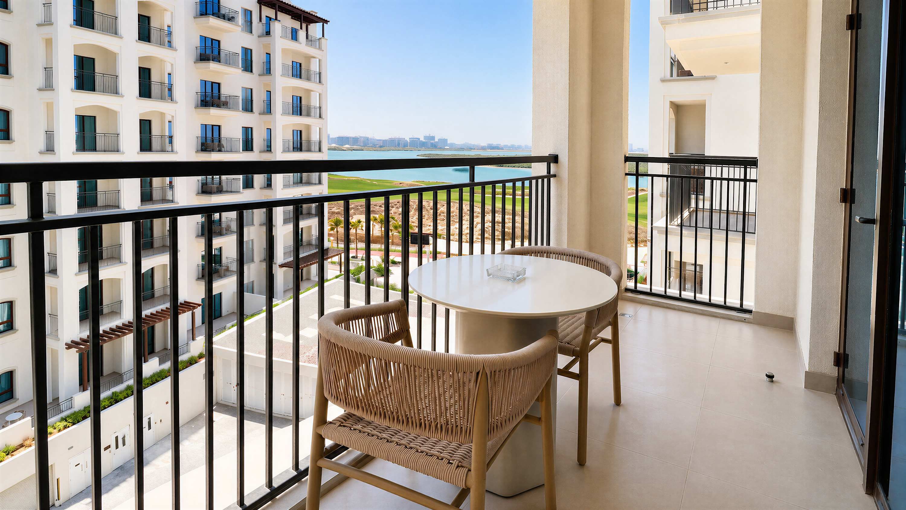 A balcony with a round table and two wicker chairs, overlooking a cityscape and a body of water.