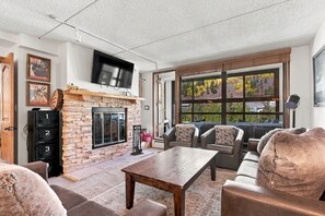 Ein Wohnzimmer mit Kamin aus Stein, einem darüber montierten Flachbildfernseher und einem großen Fenster mit Blick auf Bäume.