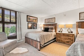 Ein Schlafzimmer mit einem großen Bett, zwei Nachttischen mit Lampen, einem Sessel und Blick ins Freie durch ein Fenster mit Jalousien.