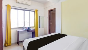 A hotel room with a bed, a desk, a window with curtains, an air conditioning unit, and a door. - Hotel O Rane Nagar (Nashik)