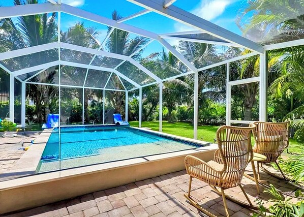 A glass-enclosed swimming pool with a slide, surrounded by wicker chairs and lush greenery.