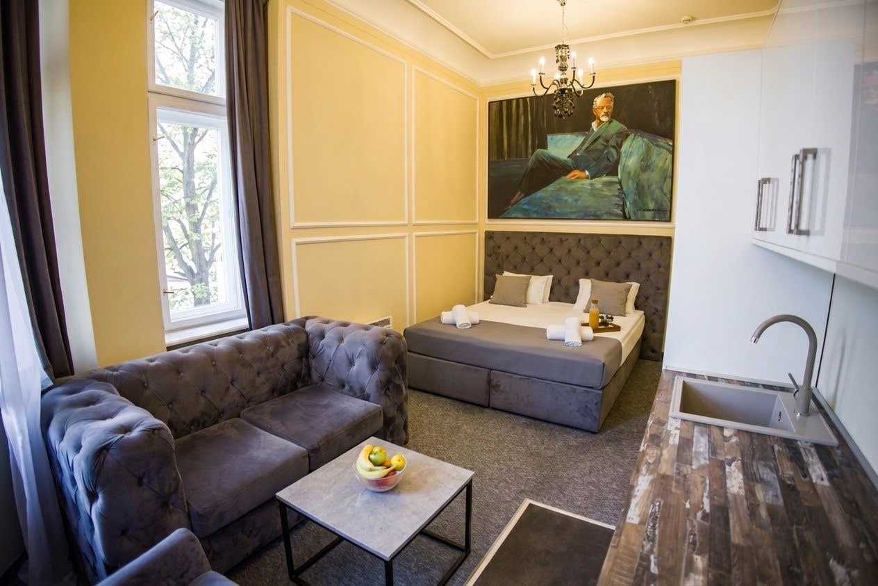 Une chambre d’hôtel compacte comprenant un canapé, une petite table avec des fruits, un lit avec des oreillers, un tableau et un lavabo.